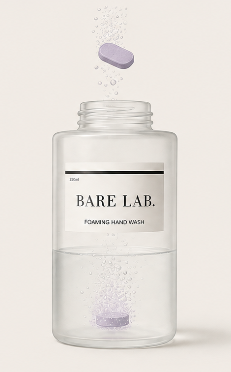 A clear cylindrical bottle labeled 'BARE LAB. FOAMING HAND WASH' on a light beige background, with a lavender oval tablet floating above the open top and another tablet sinking into the water inside.